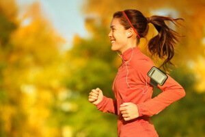 Woman runner running in fall autumn forest listening to music on smartphone using earphones. Female fitness girl jogging on path in amazing fall foliage landscape nature outside.
